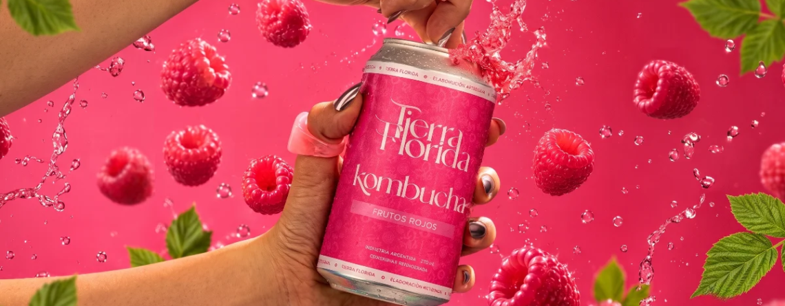 Mano abriendo una lata fría de Kombucha de Frutos Rojos de Tierra Florida, rodeada de frambuesas frescas sobre fondo rosa.