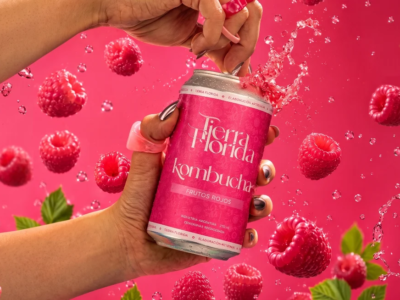 Mano abriendo una lata fría de Kombucha de Frutos Rojos de Tierra Florida, rodeada de frambuesas frescas sobre fondo rosa.