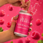 Mano abriendo una lata fría de Kombucha de Frutos Rojos de Tierra Florida, rodeada de frambuesas frescas sobre fondo rosa.